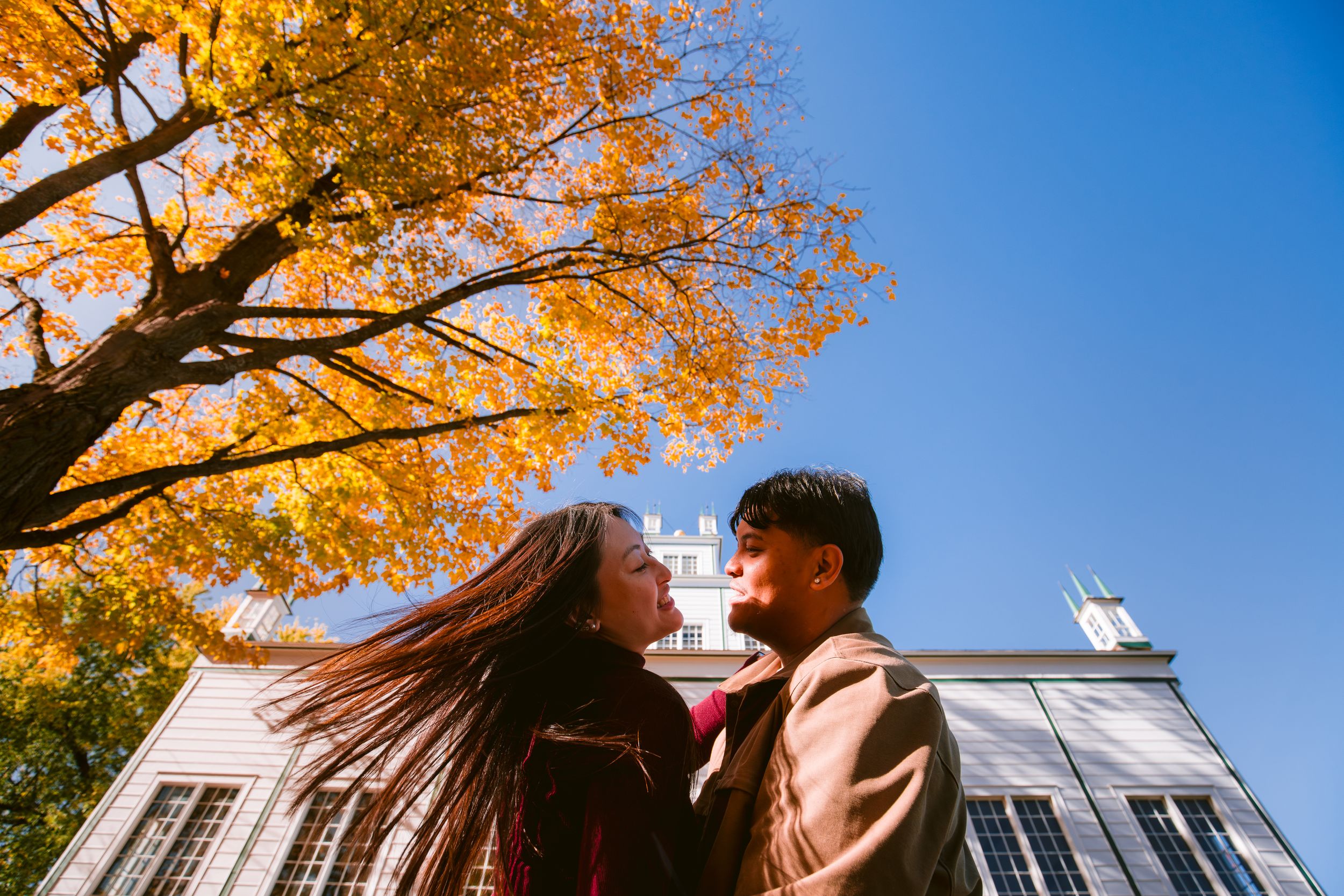 A couple shares an intimate moment beneath golden autumn trees with a white building in the background.