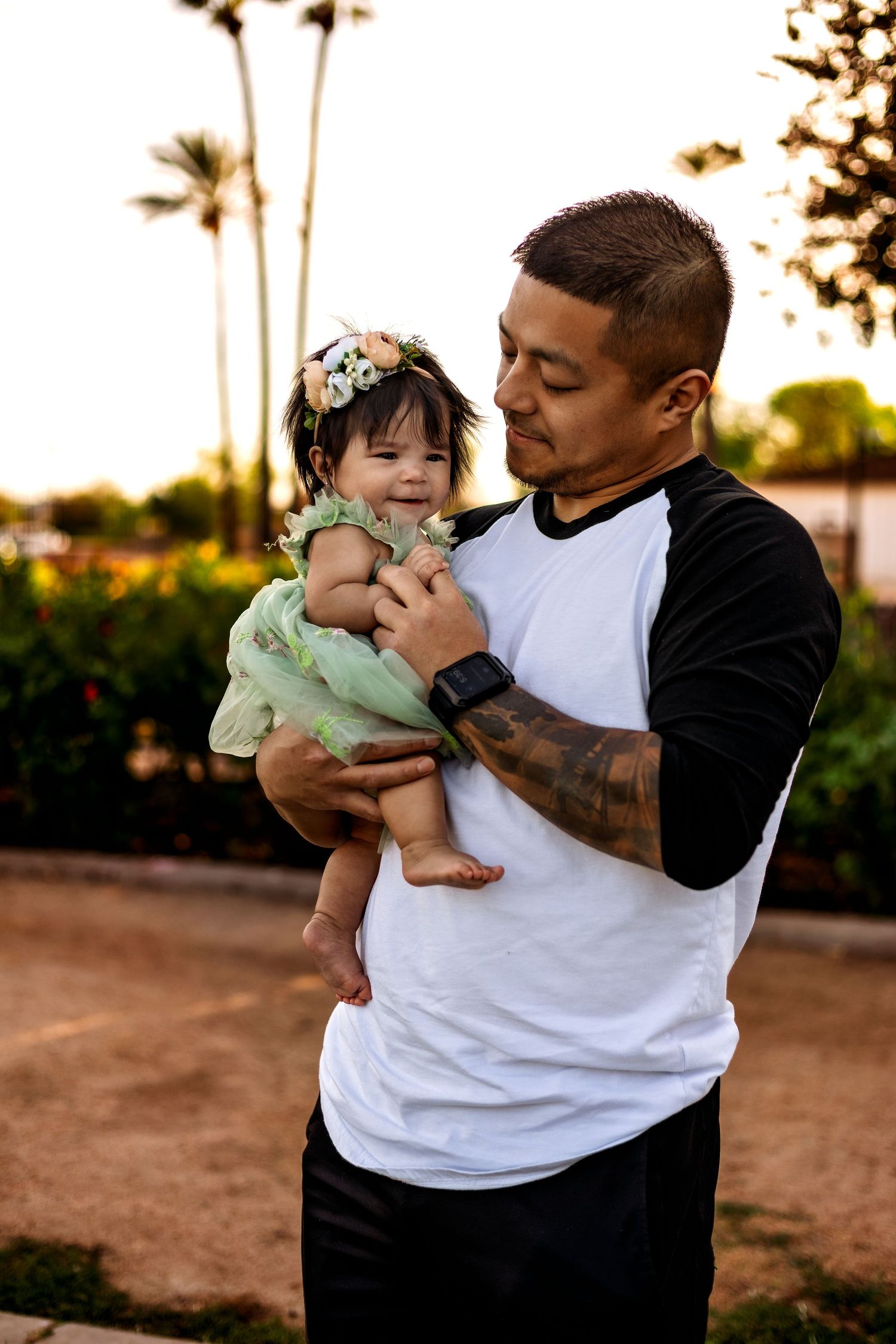 A parent and baby share an intimate moment outdoors at sunset with palm trees visible in the background.