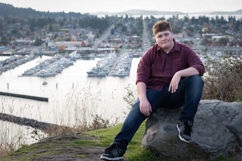 A person sits casually on a rock overlooking a scenic marina with boats and mountains in the background.