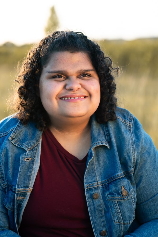 Someone wearing a denim jacket and burgundy shirt smiles warmly during golden hour outdoors.