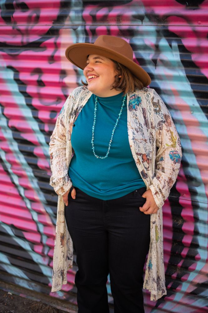 Person in brown hat and floral kimono poses happily against colorful graffiti wall.
