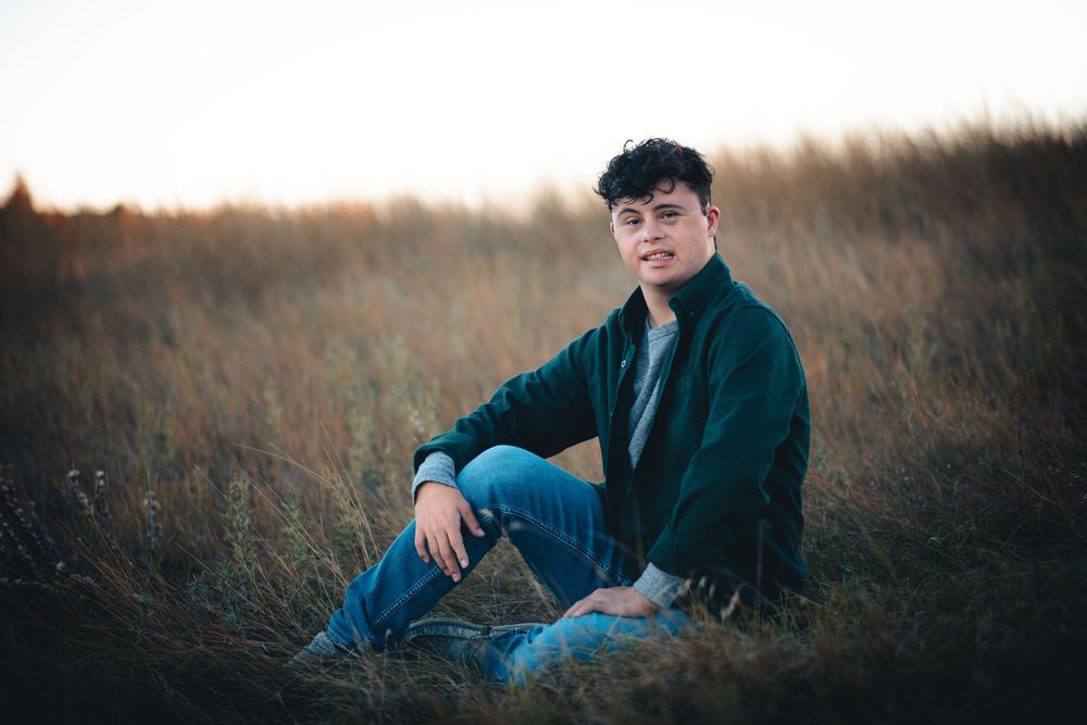 Young person in navy jacket sits thoughtfully in tall grass field during sunset.