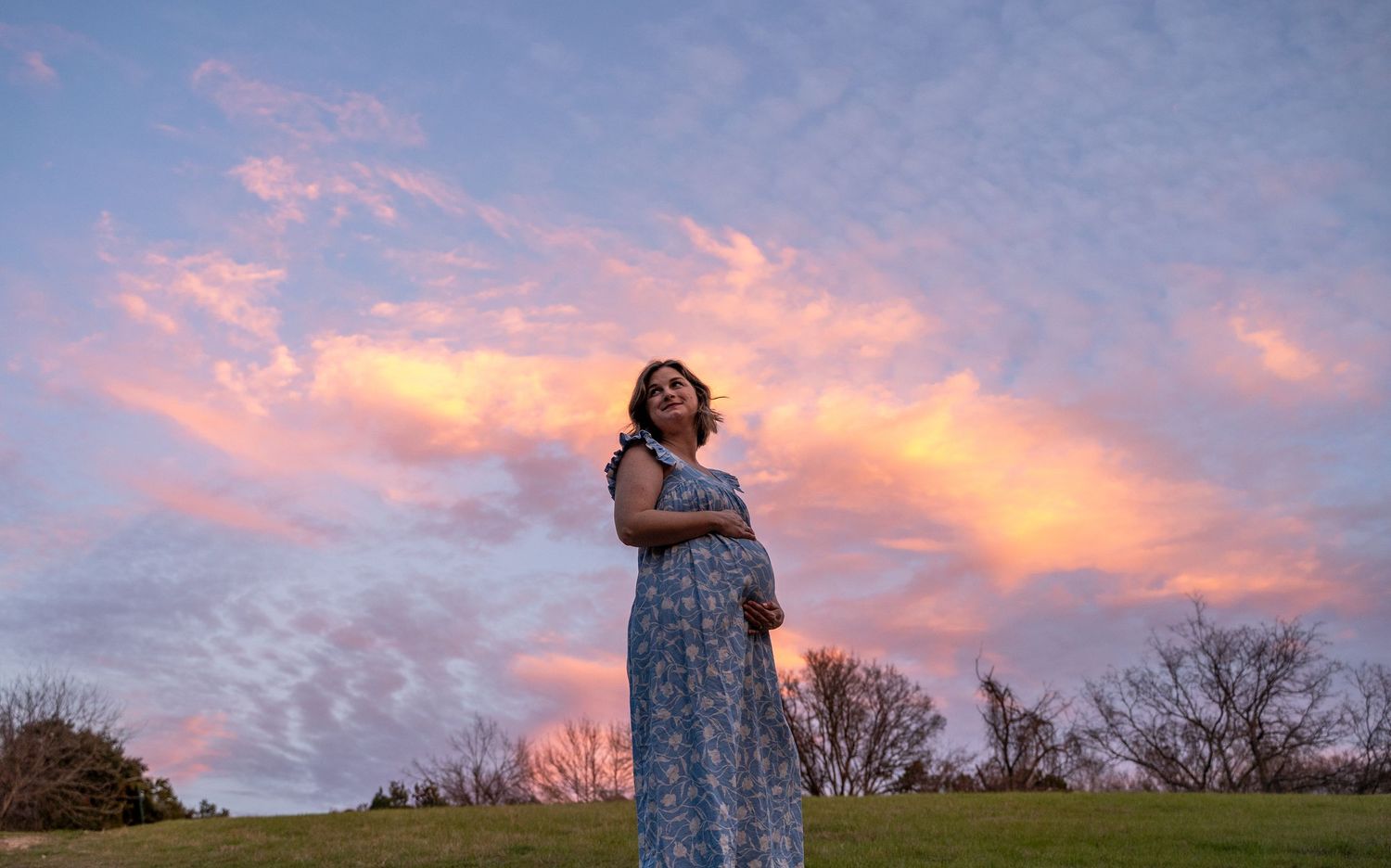Person laying on wooden planks outdoors in a blue patterned dress during sunset with trees in background.