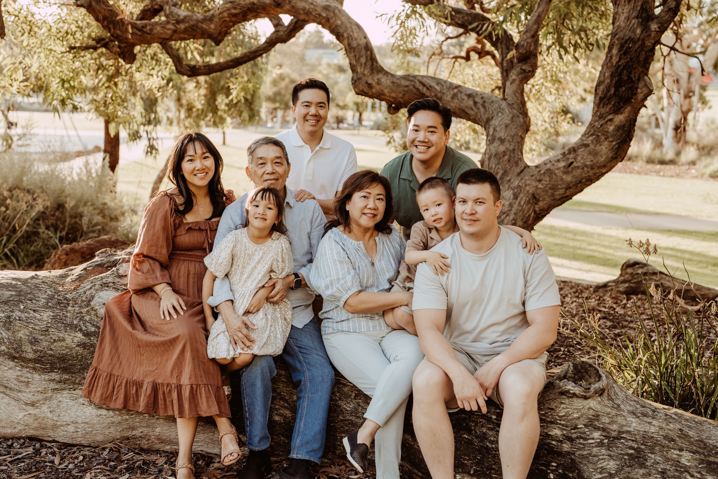 Extended family gathering outdoors under a sprawling tree on a sunny day.