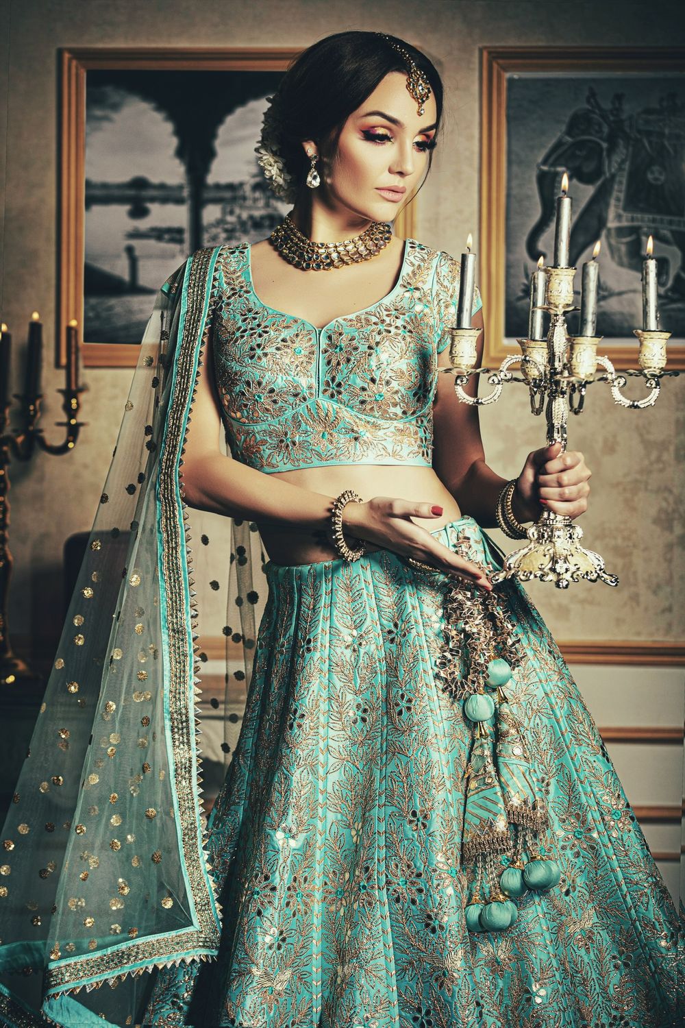 A bride in a teal embroidered lehenga holds a silver candelabra, adorned with gold jewelry in an elegant interior.