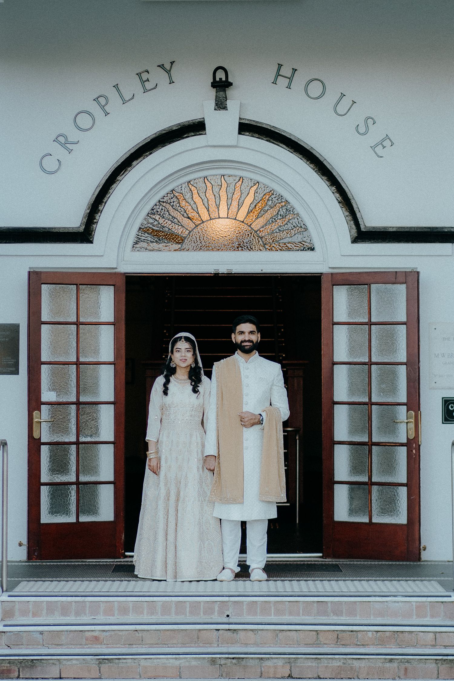 A couple stands in formal attire at the elegant entrance of Cropley House featuring an ornate fanlight and white columns.
