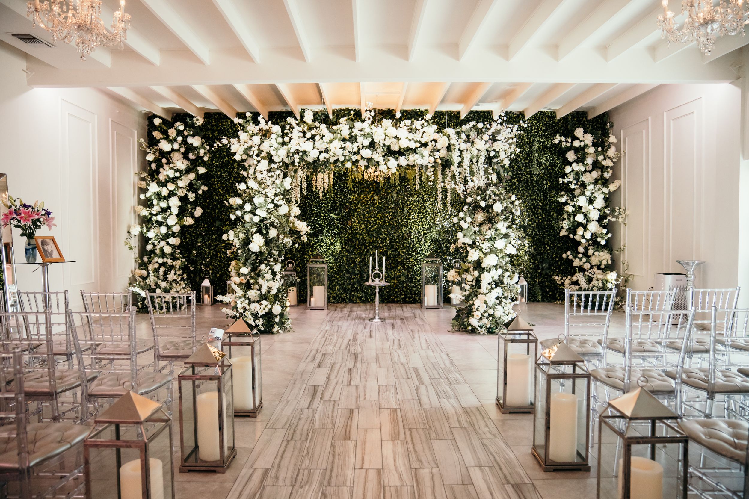 Indoor wedding ceremony space with white floral arch and lantern-lined aisle featuring rustic wooden flooring.