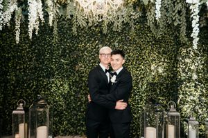 Couple embraces under romantic garden setting with hanging lights and greenery wall backdrop.