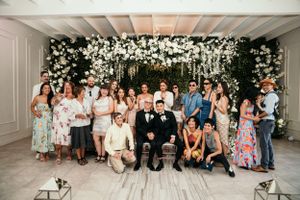 A wedding party gathers under an elegant white floral arch against a light gray wall for a group photo.