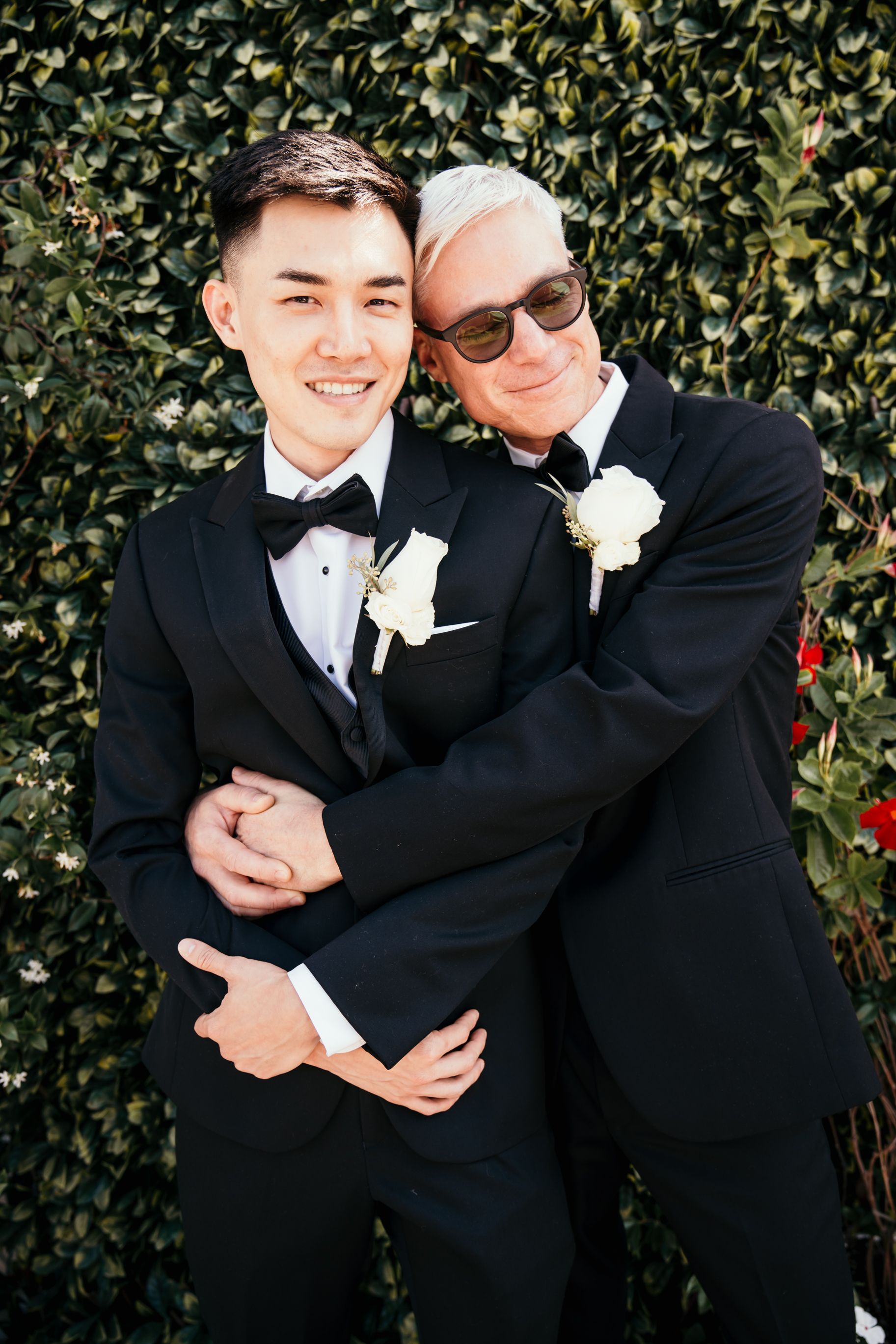 Two people in black tuxedos and white boutonnieres embrace while posing against a leafy hedge backdrop.