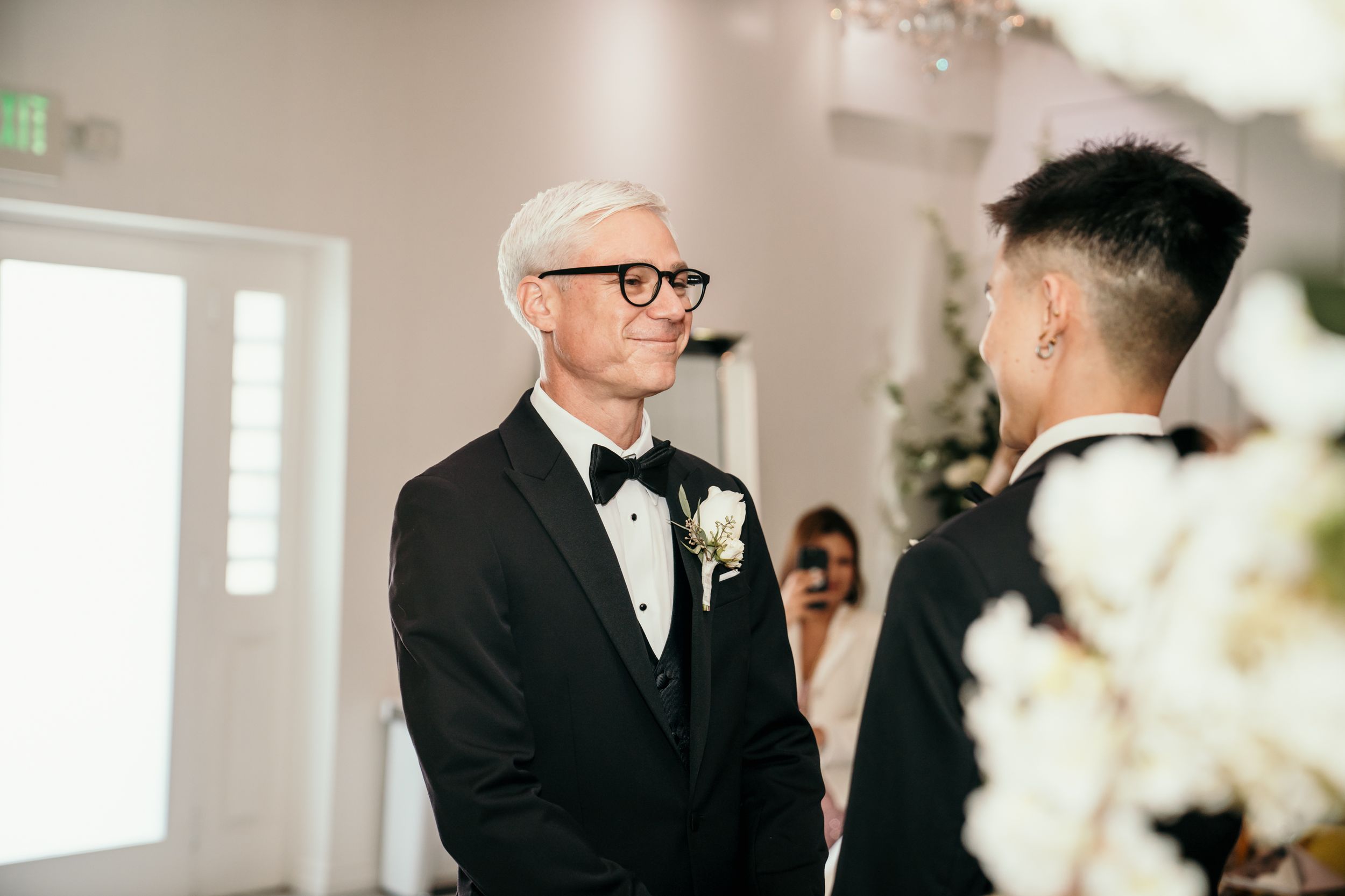 Two people in black tuxedos and bow ties share an intimate moment during a wedding ceremony.