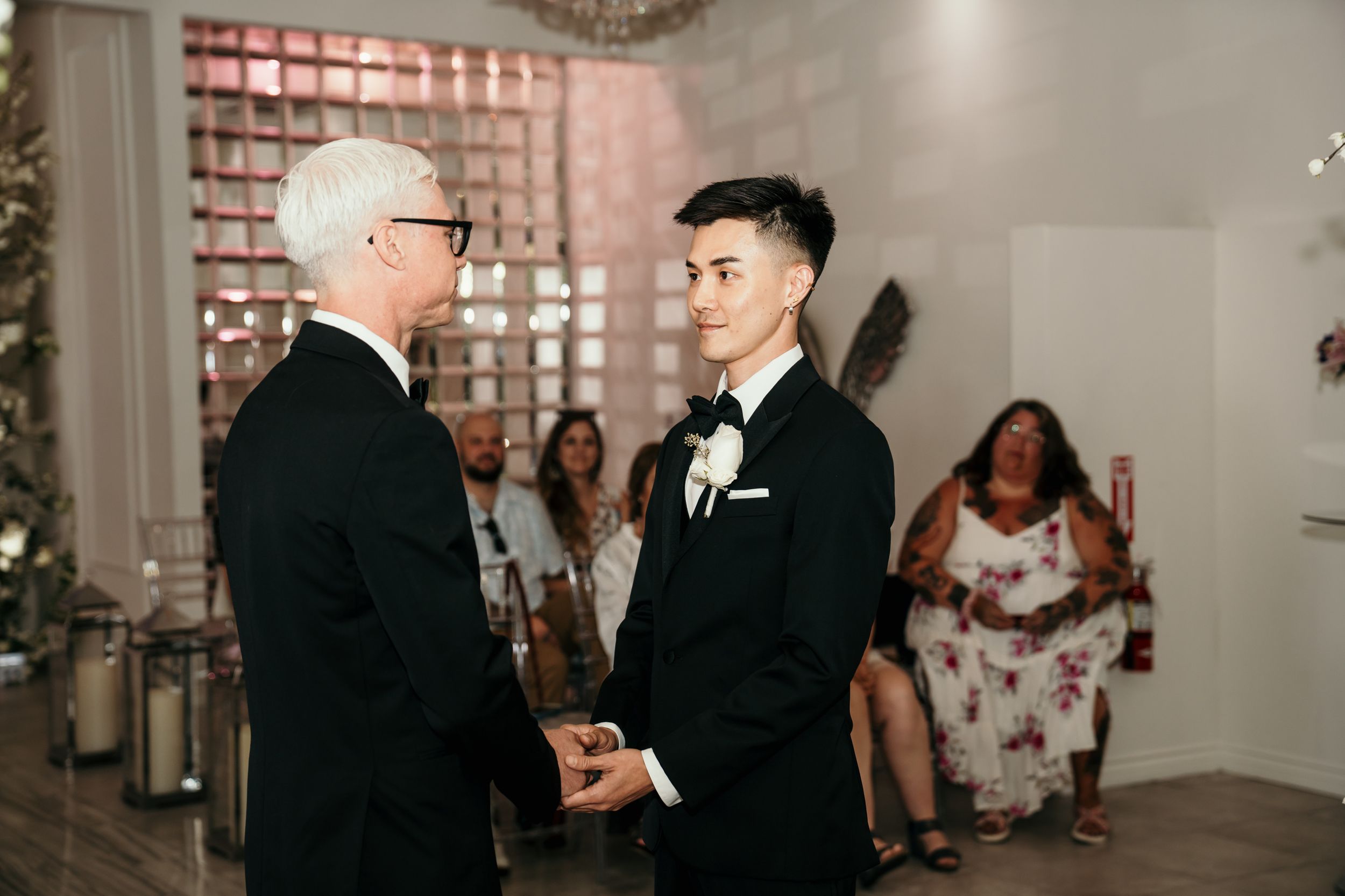 Two people in black suits stand facing each other during an indoor wedding ceremony with guests in background.
