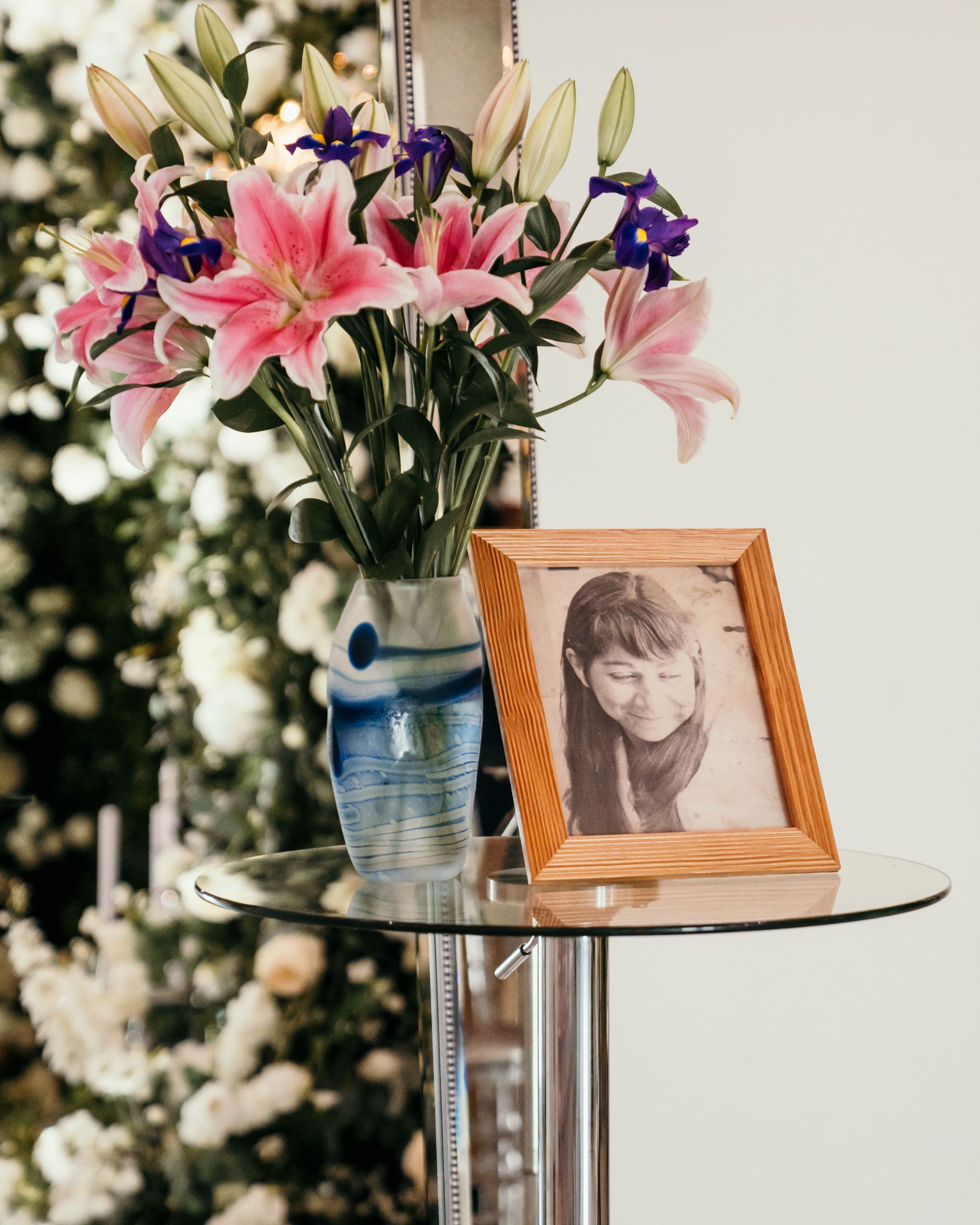 A blue vase with pink lilies and purple flowers sits on a glass table next to a wooden picture frame.