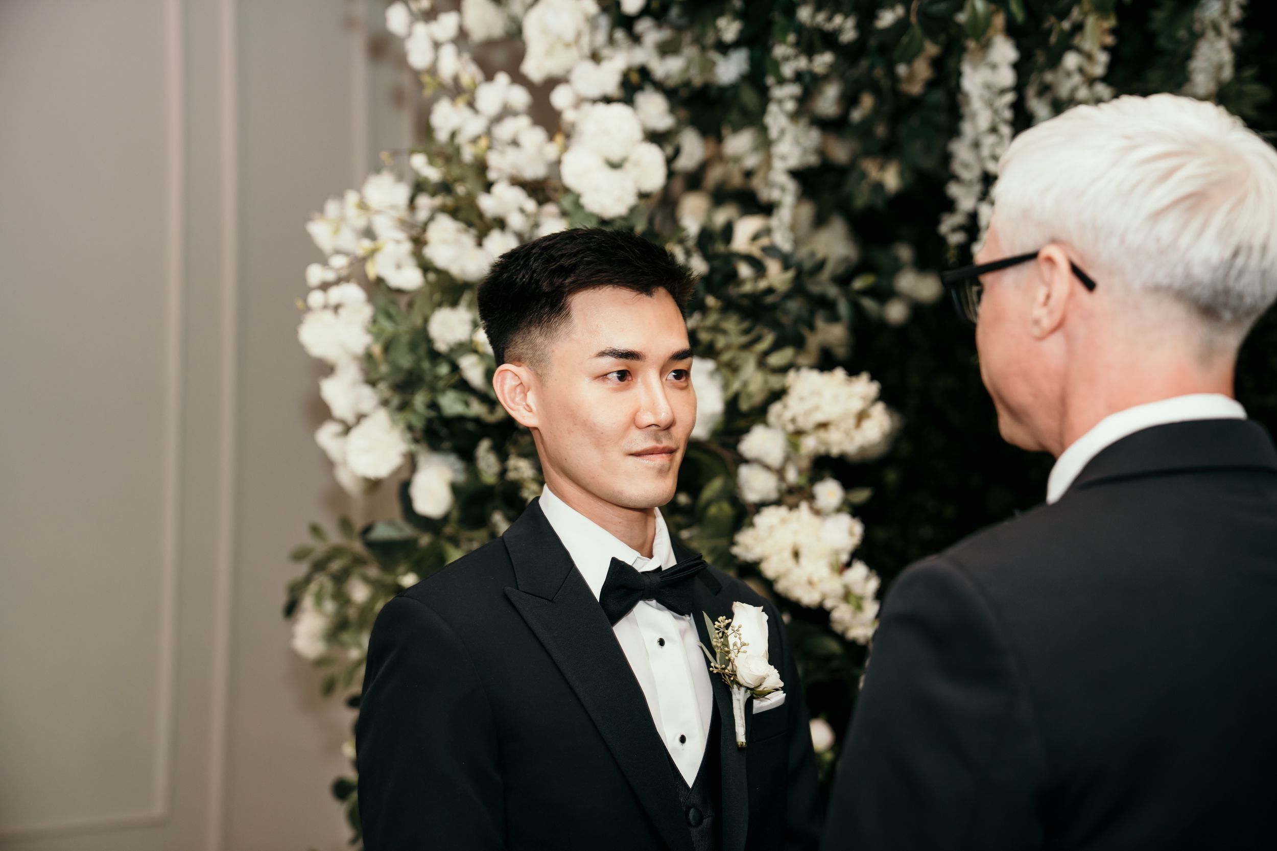 Close up shot at a formal event with two people in suits conversing in front of an elegant white floral backdrop.