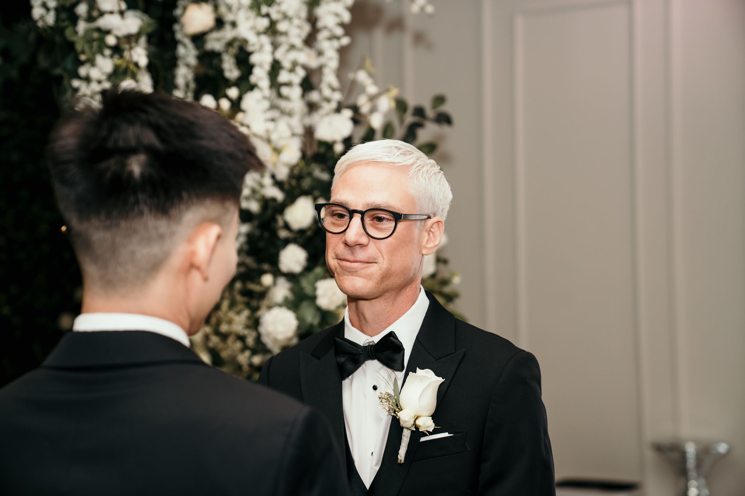 Two people in formal black tie wedding attire are captured during an elegant moment with white floral decor in background.