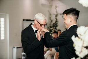 An emotional moment between two people in formal black suits during an indoor event surrounded by white floral decor.