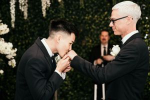 A touching moment at a wedding ceremony with two people in black suits against a greenery backdrop.