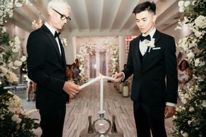 Two people in black suits perform a wedding ceremony with decorative floral arches and candles in an elegant indoor setting.