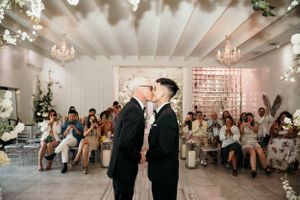 Couple shares intimate moment during wedding ceremony in elegantly decorated white room with crystal chandeliers.