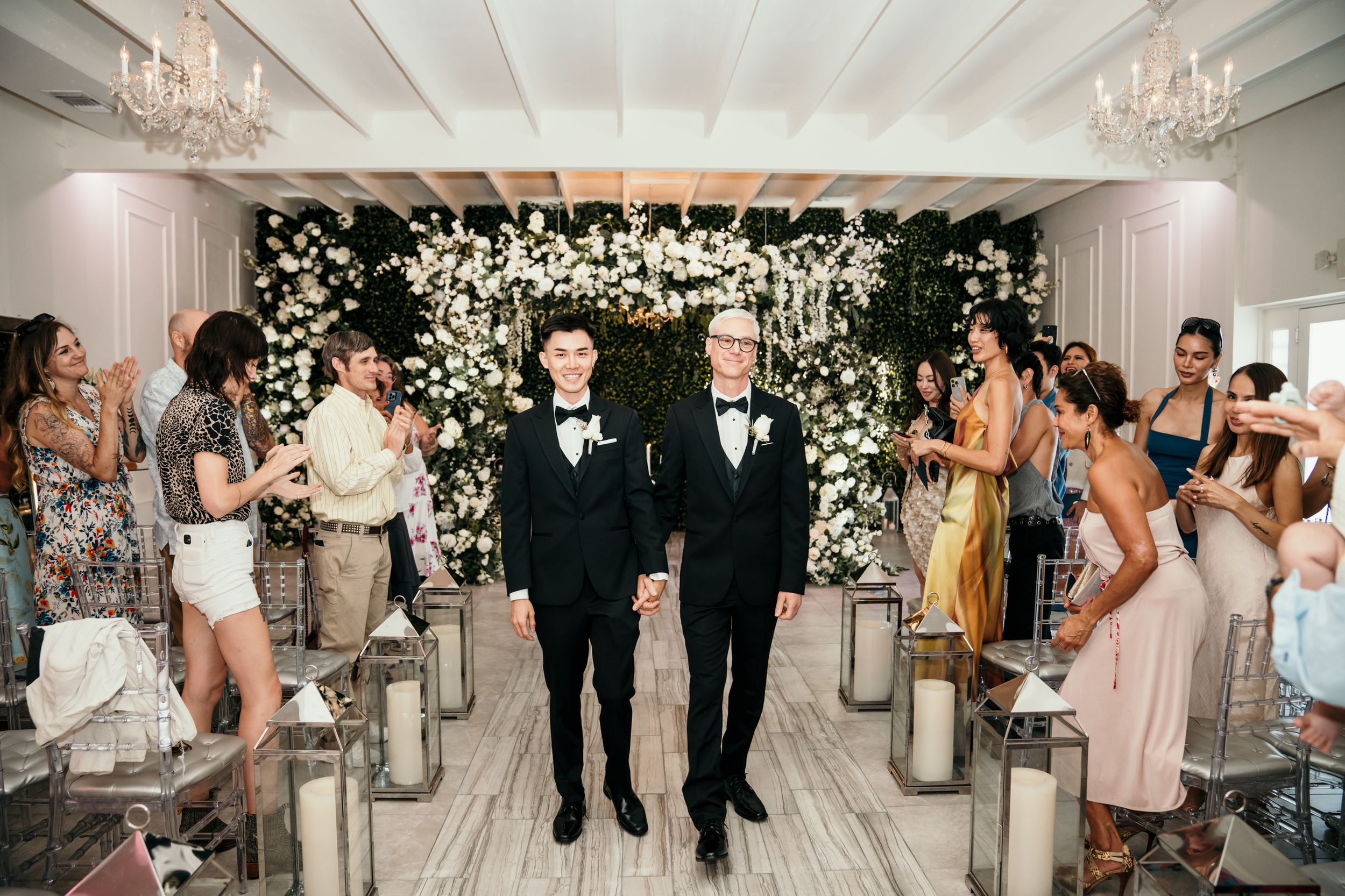 A romantic wedding ceremony setup with a white floral arch backdrop and lantern-lined aisle in an elegant indoor venue.