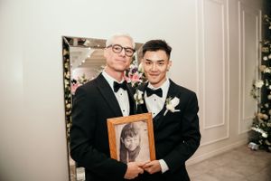 Two people in black tuxedos stand together while holding a framed photograph at a formal event.