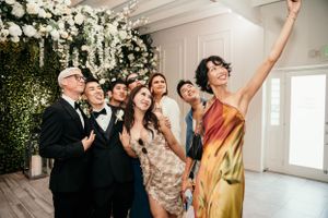 Wedding party group posing joyfully together against a floral wall display at a celebration.