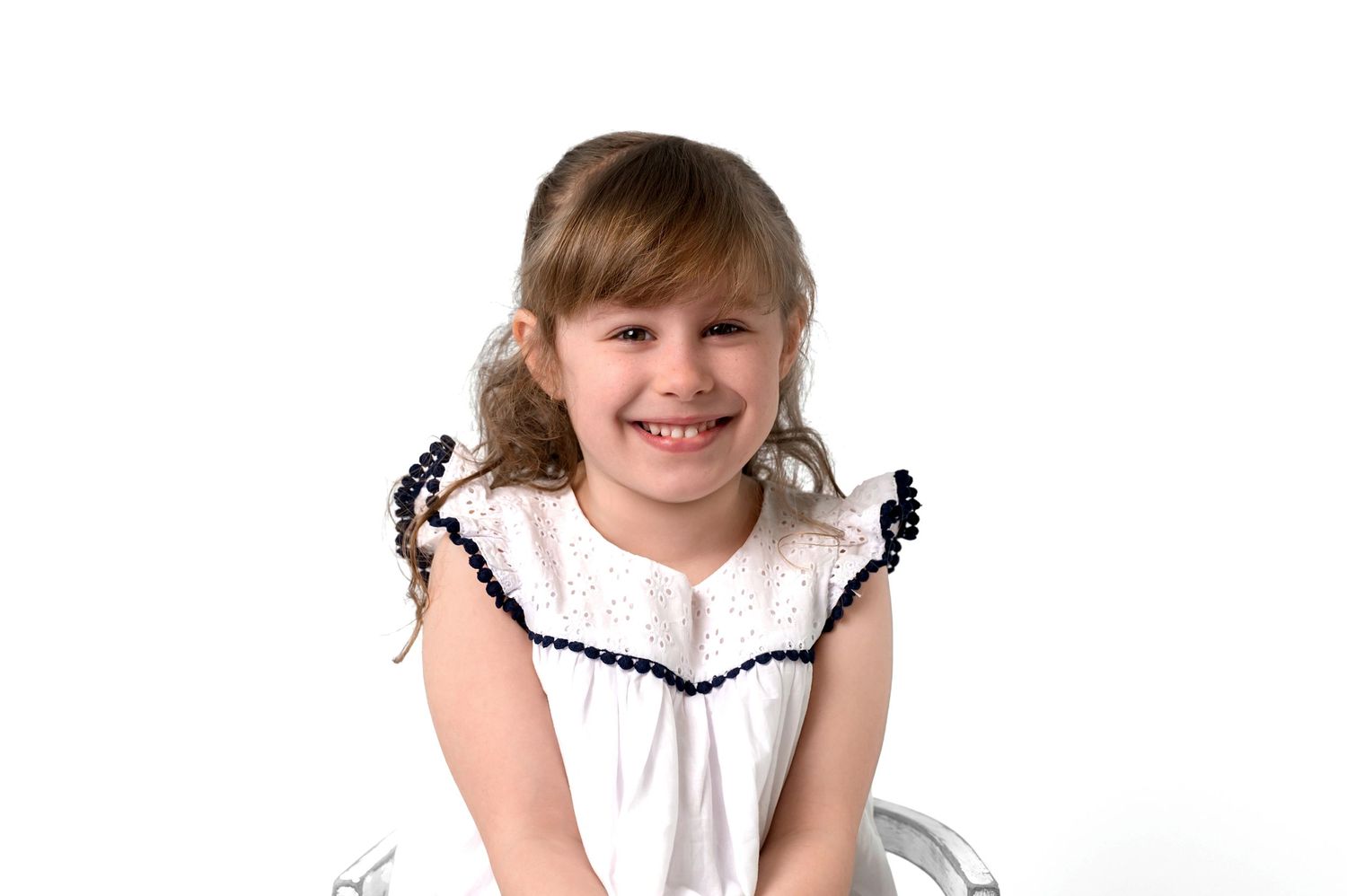 A young child wearing a white dress with black trim around the sleeves sits while smiling against a white background.
