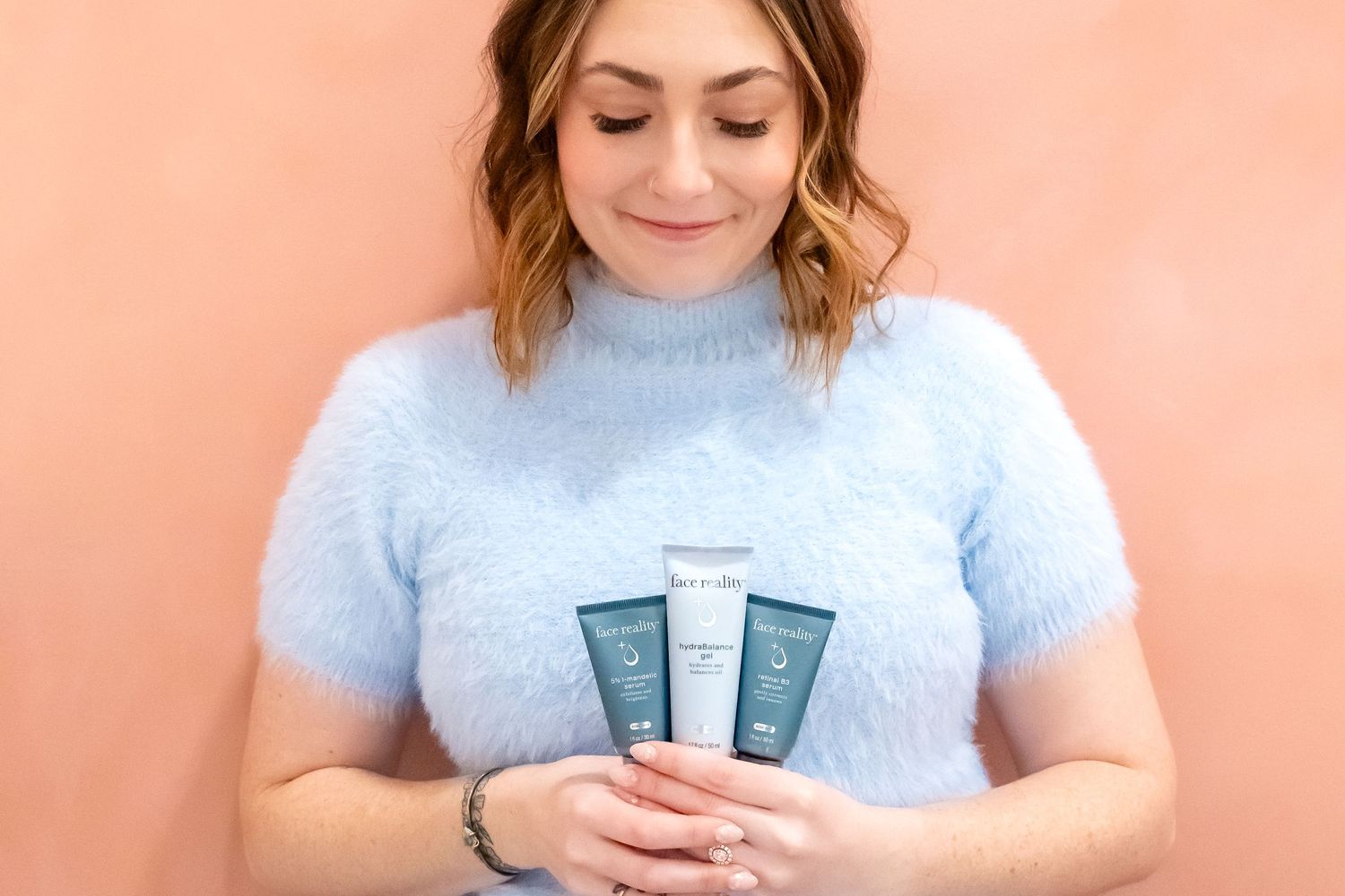 Woman wearing a soft blue fuzzy sweater holds three skin care products against a peach colored backdrop.