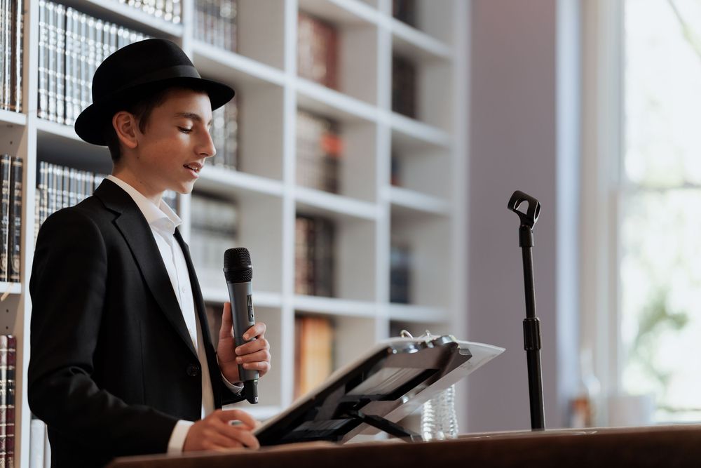 A speaker in a black suit and hat stands at a podium with microphone in a bright room with bookshelves.