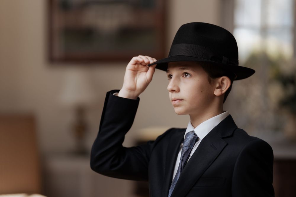 Young person in a dark suit and hat poses formally against an interior background.