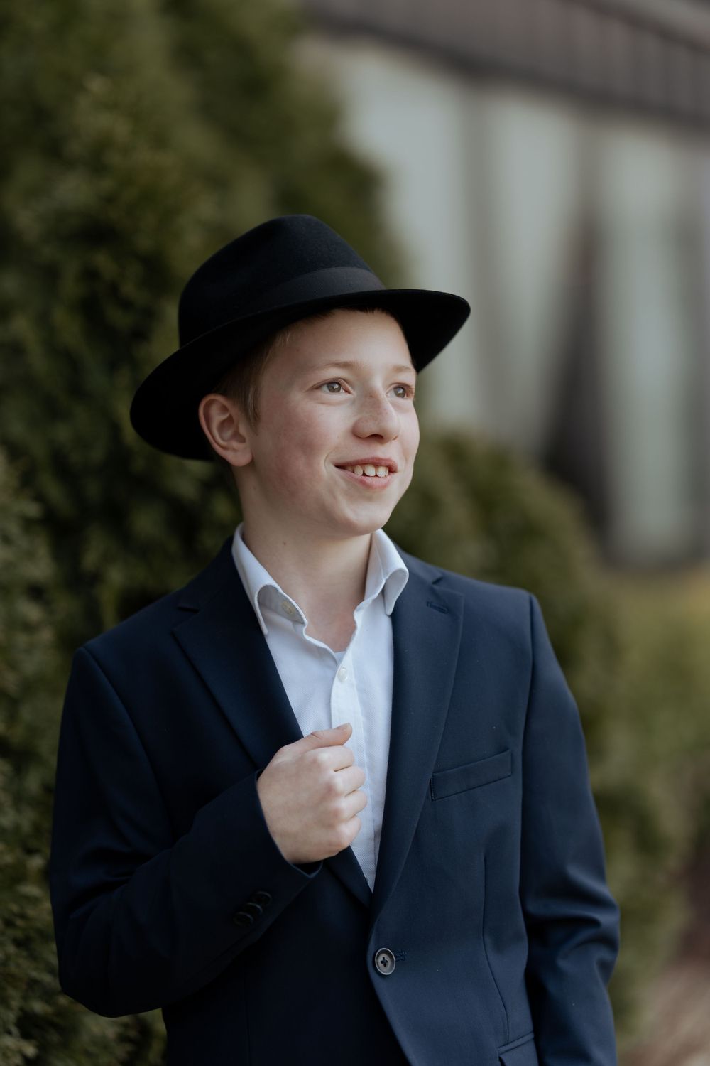 Young person in navy suit and black hat smiles while standing against green hedge backdrop.