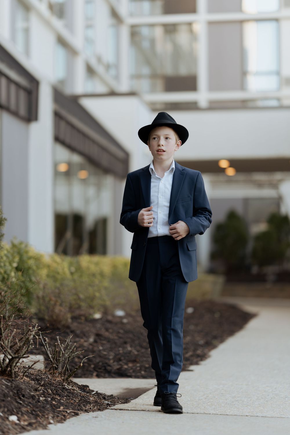 Young person in formal navy suit and black hat walks along building pathway.