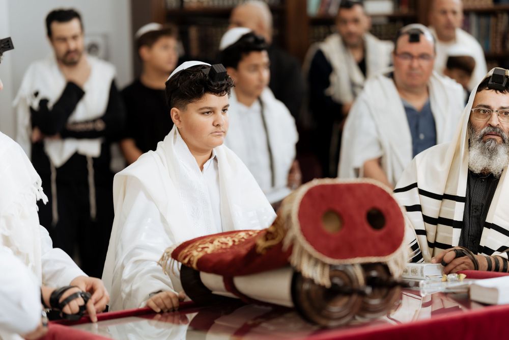 People gather at a synagogue table with religious texts and prayer shawls for a traditional ceremony.