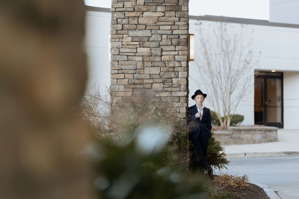 Person in dark suit and hat standing near stone wall and landscaping.