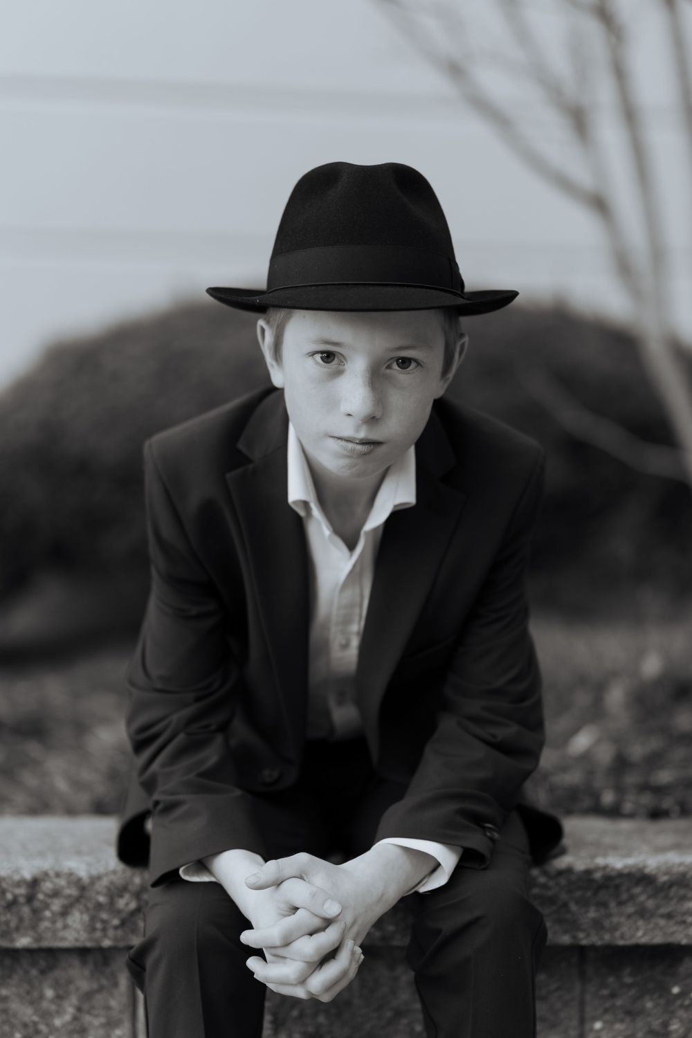 A young person in formal attire and fedora hat sits contemplatively on steps in black and white photography.