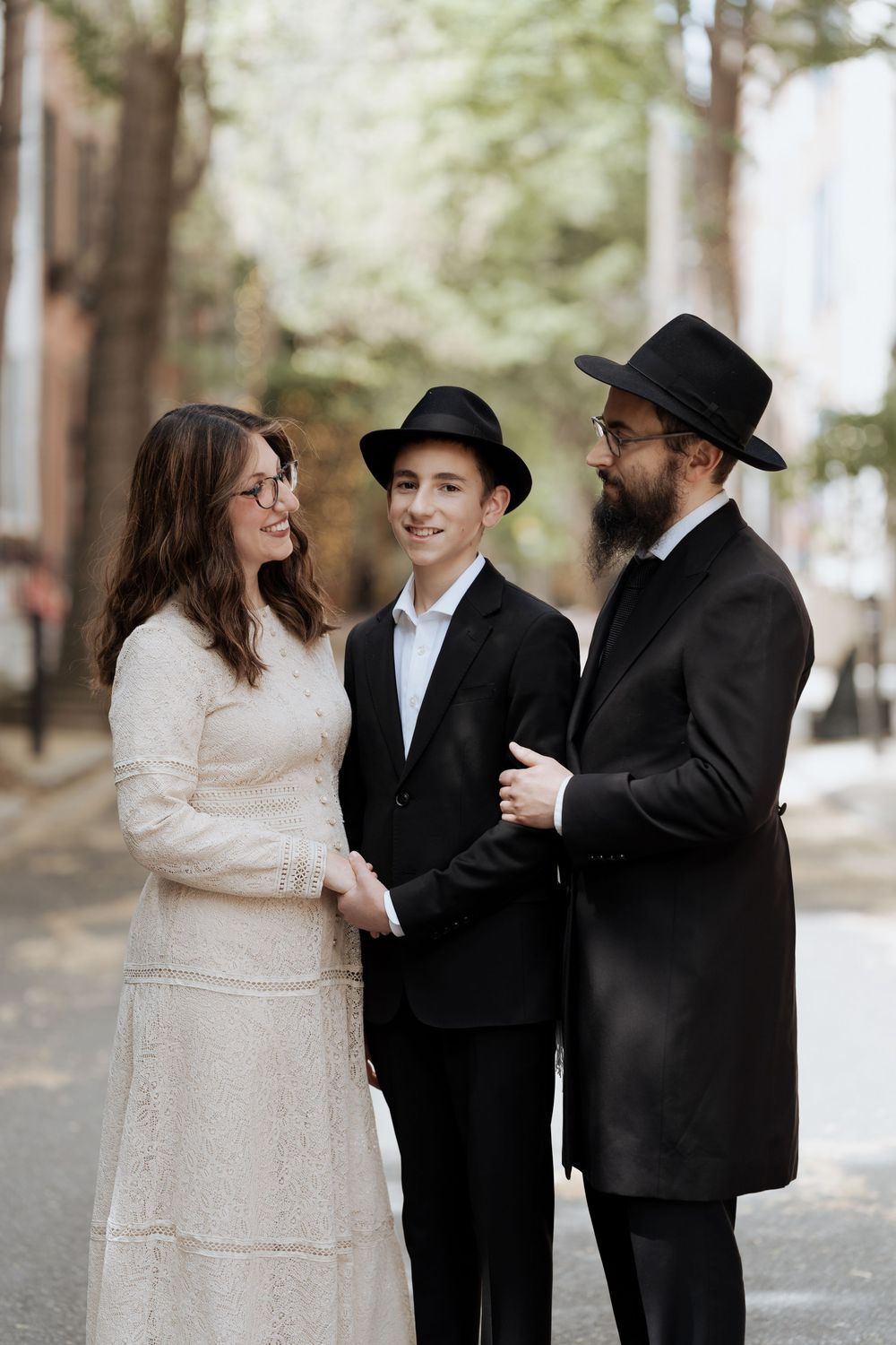 A group stands together on a tree-lined street wearing formal Jewish Orthodox attire including black hats and a cream dress.