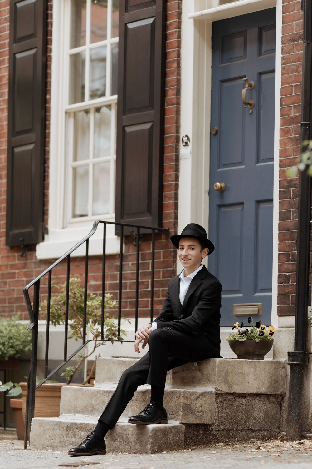 A person in formal attire sits on brick steps of a historic townhouse with a blue door.
