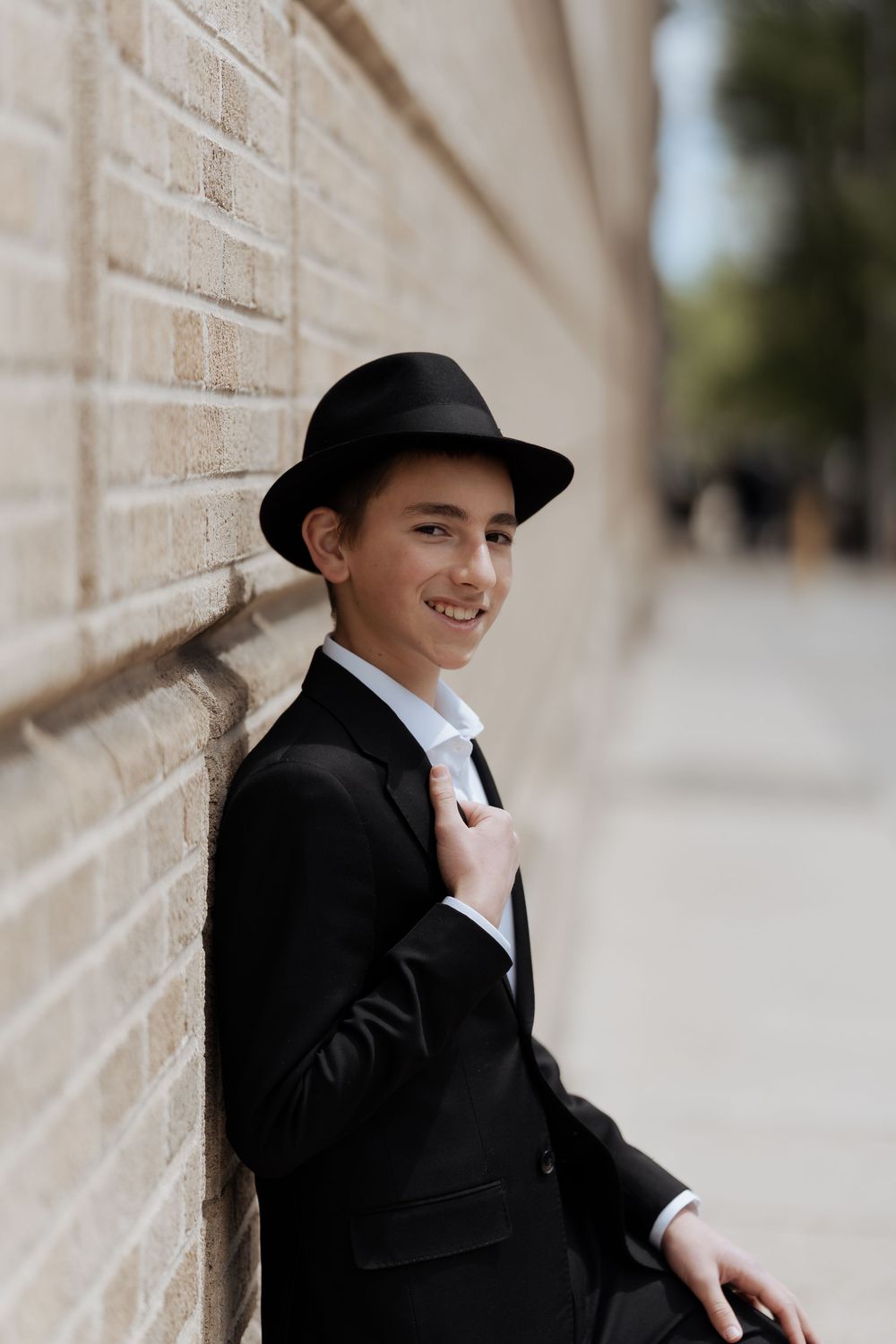 A stylish young person in a black suit and fedora leans against a beige brick wall with a warm smile.
