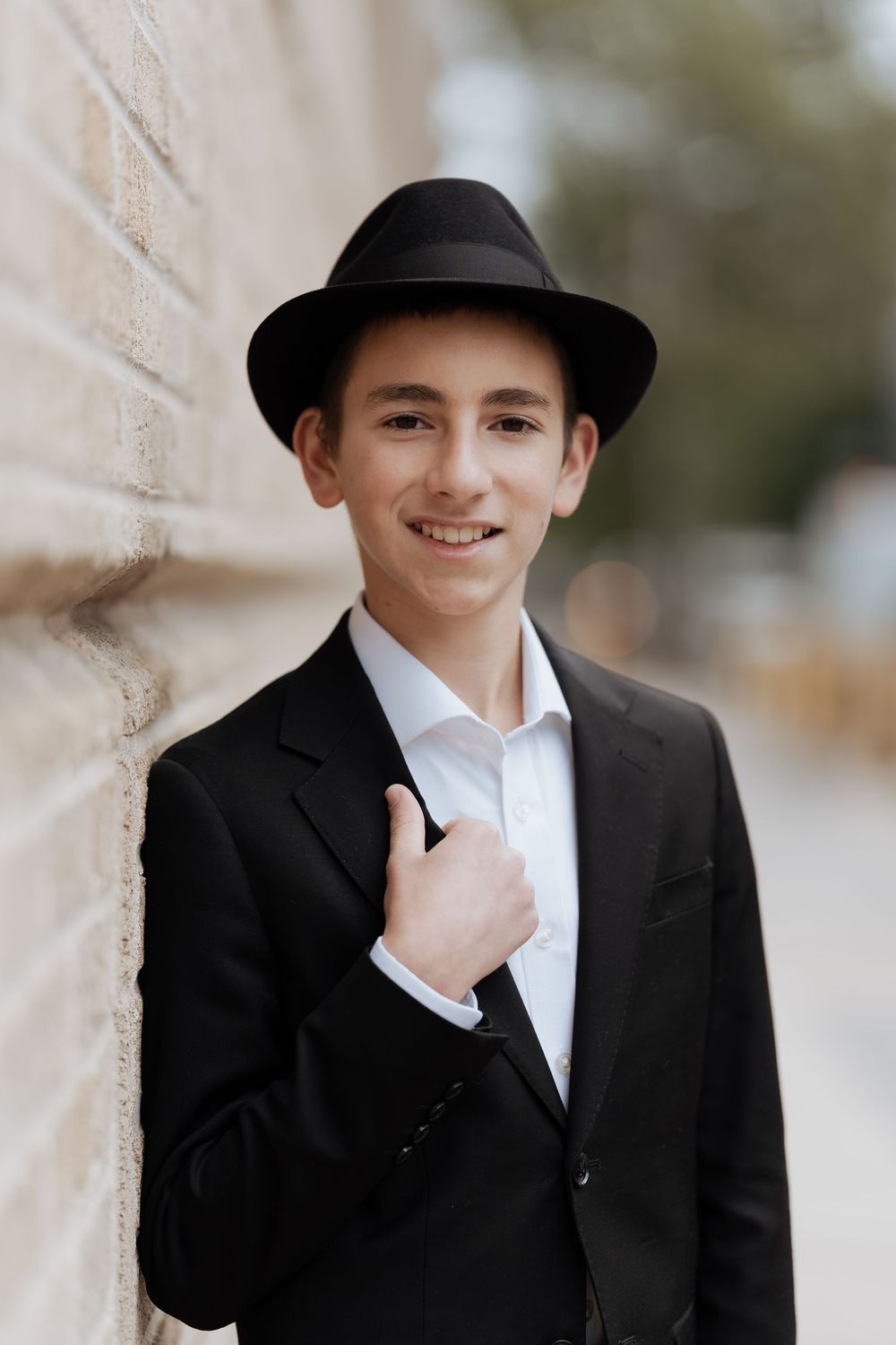 Portrait of a young person in a black suit and fedora hat leaning against a wall outdoors.