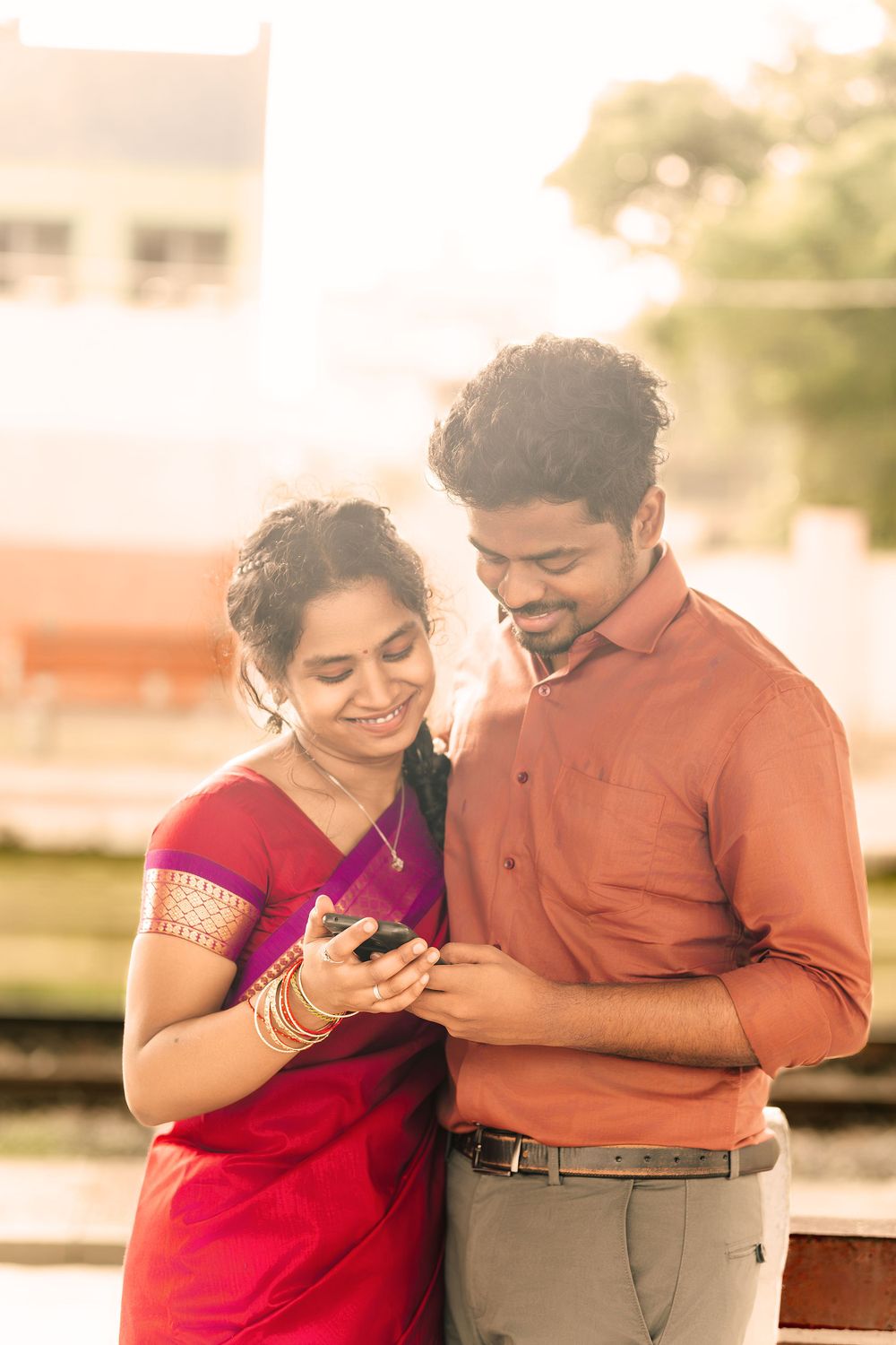 Young couple in Indian attire share a tender moment while looking at their phones together in warm sunlight.
