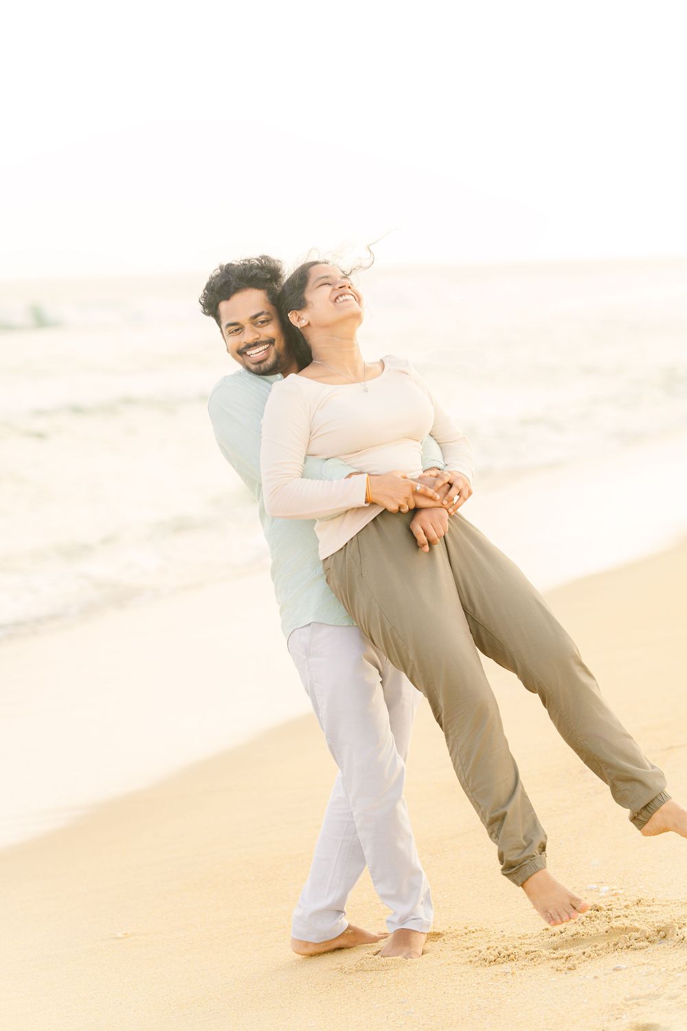 A joyful couple embraces while standing barefoot on a sunny beach at sunset.