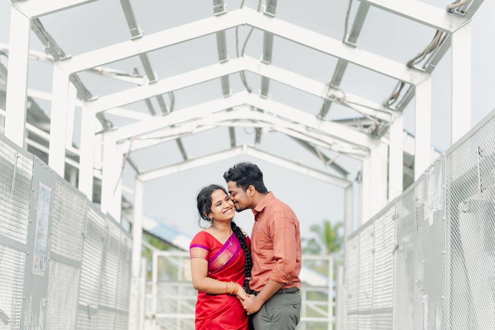 A couple in red and coral outfits embrace underneath a white covered walkway structure with architectural details.
