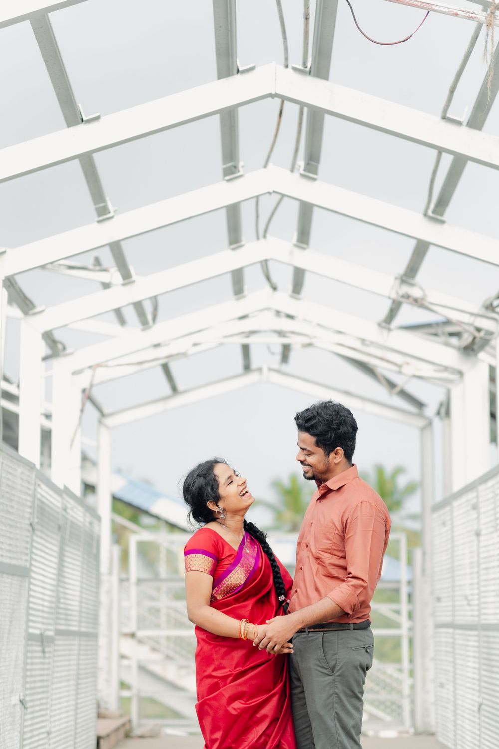 Couple in red sari and orange shirt embrace lovingly in a bright white architectural corridor.