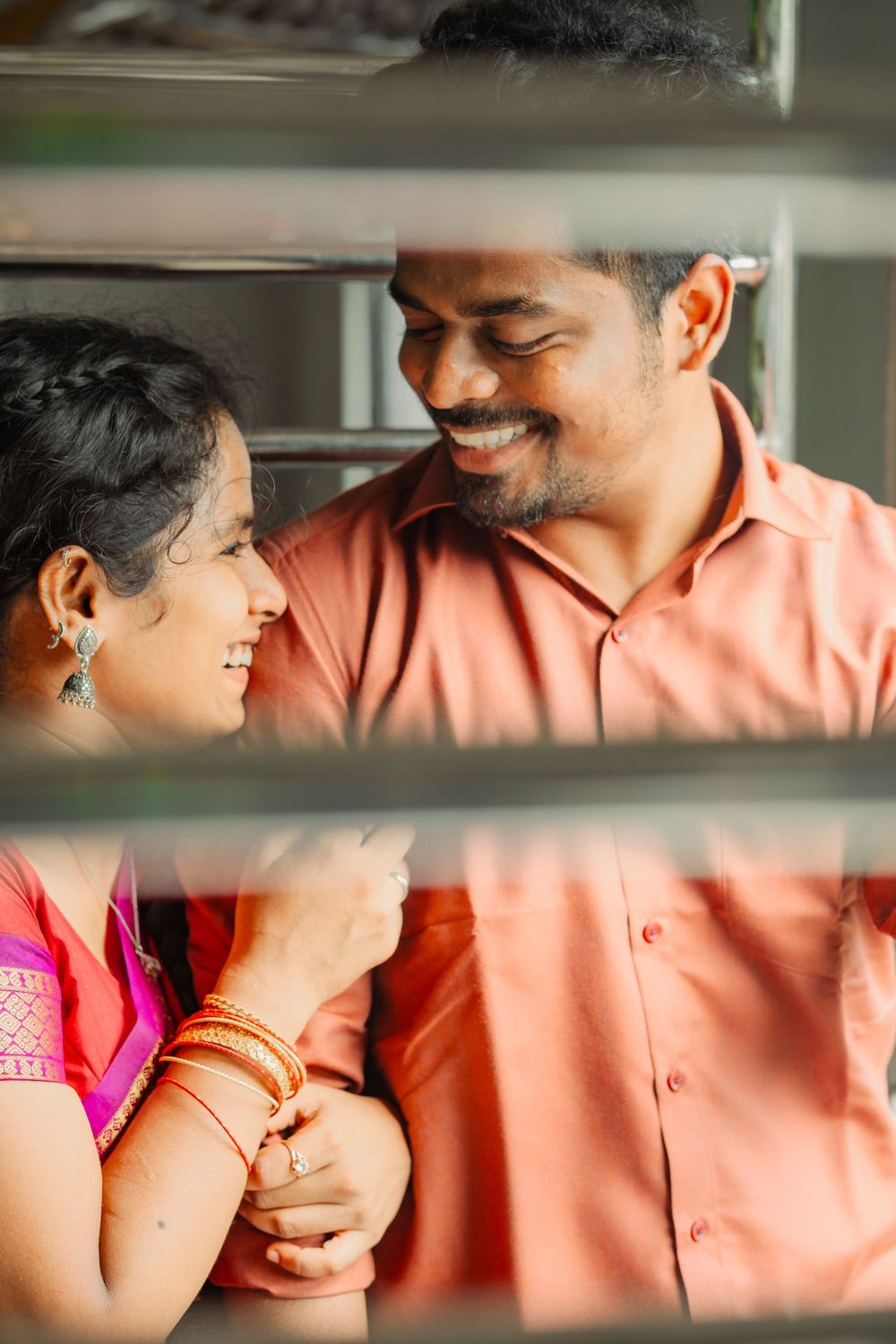 Happy couple sharing a tender moment through window blinds, wearing pink and peach colored outfits.