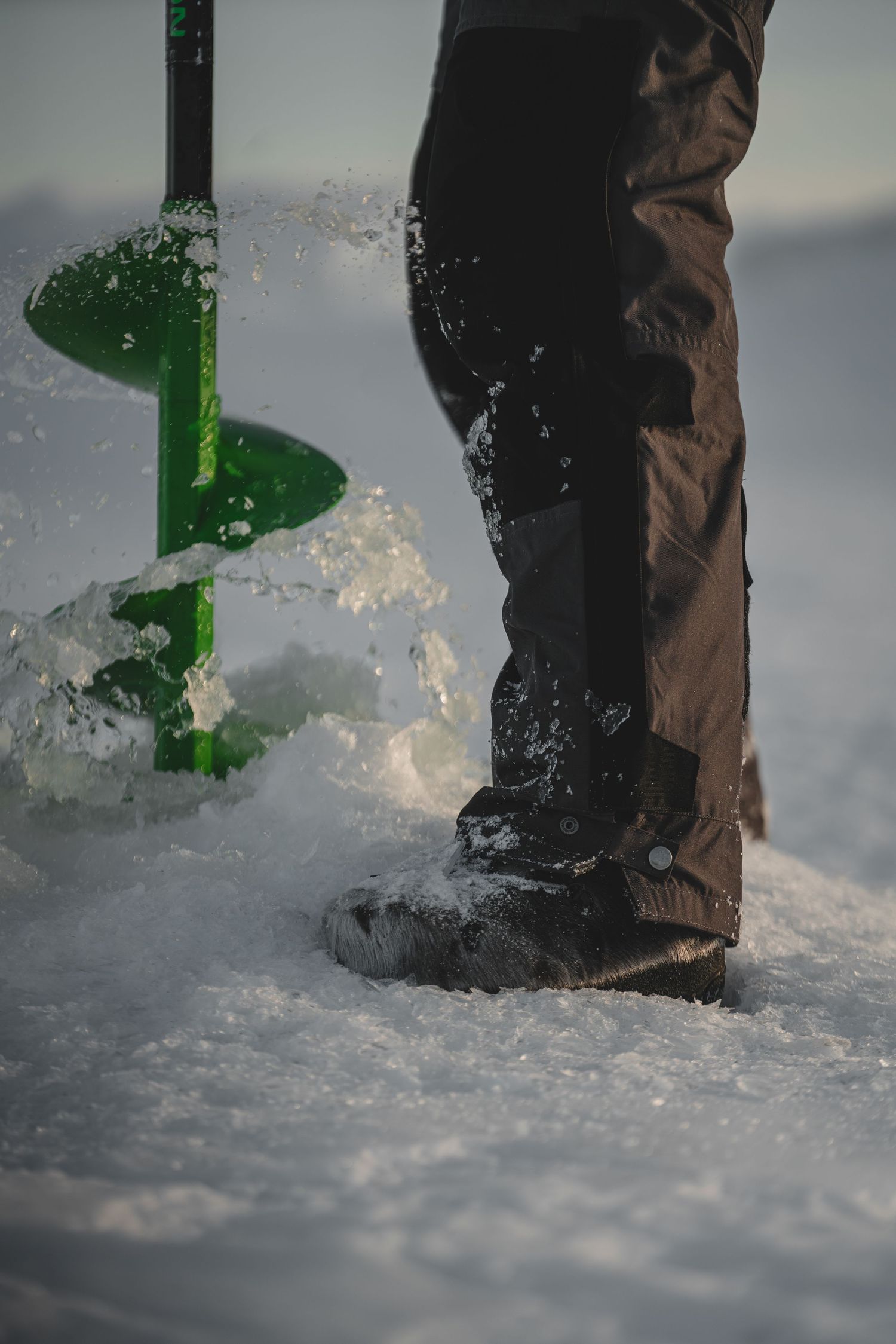 Ice auger drilling through frozen surface with snow spray.