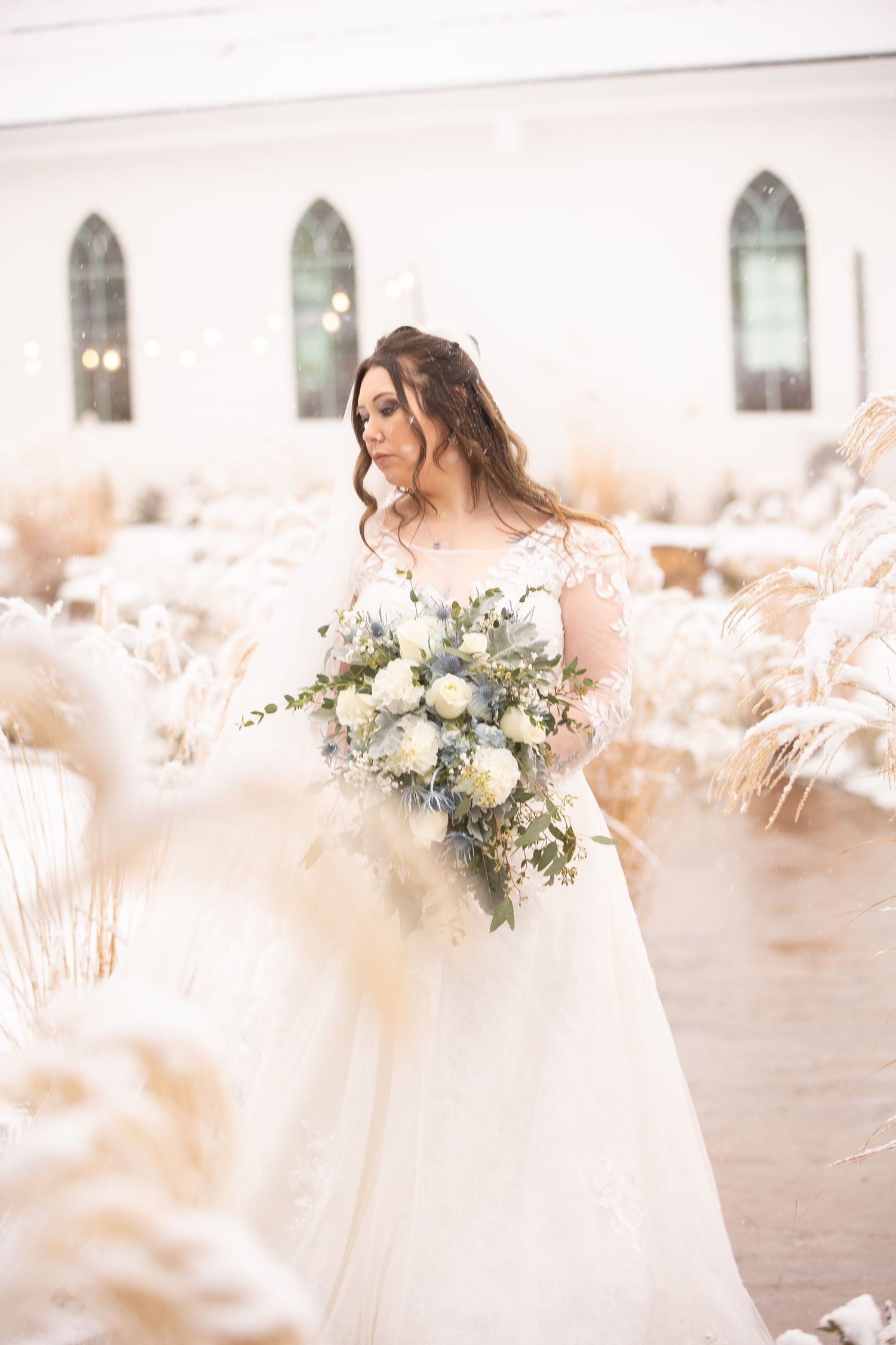 A bride in a white gown holds a bouquet while standing among dried pampas grass near a chapel.