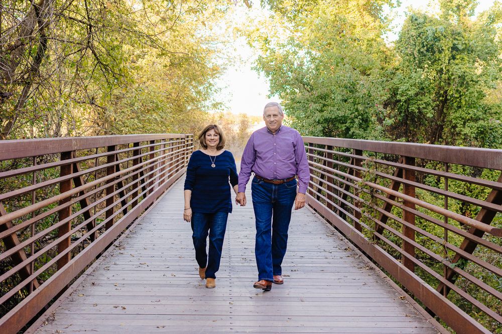 Two people walk hand in hand along a wooden bridge path surrounded by autumn trees during golden hour.
