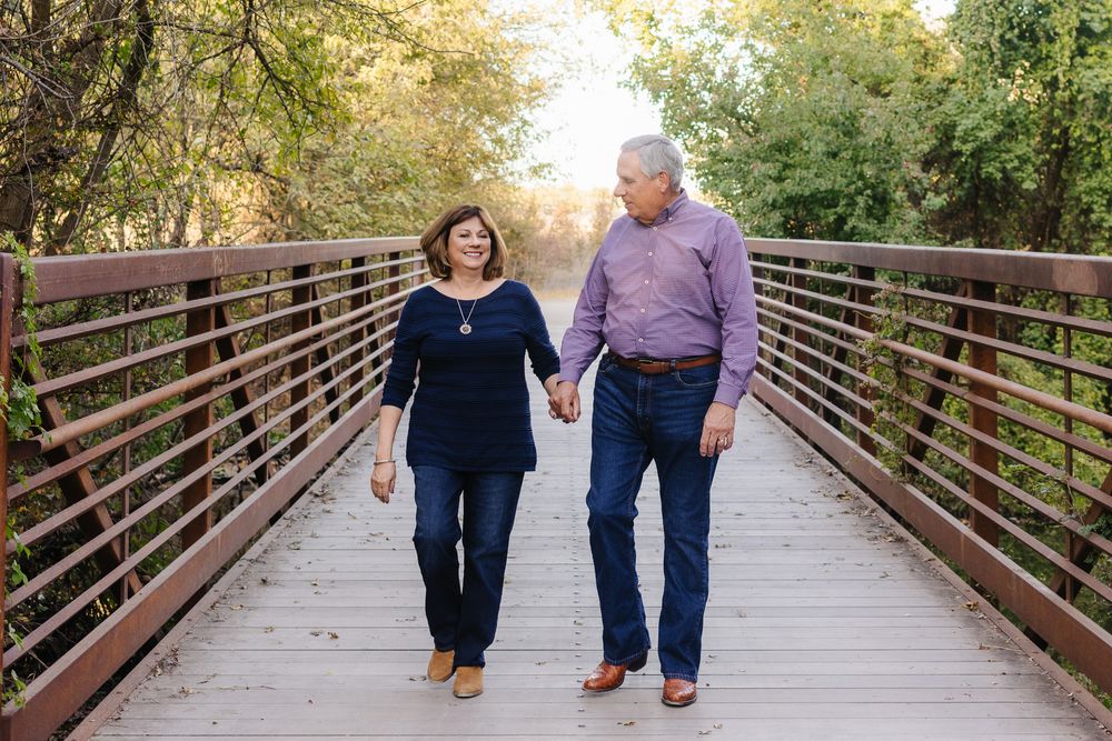 A couple walks hand in hand along a wooden bridge surrounded by autumn trees during golden hour.