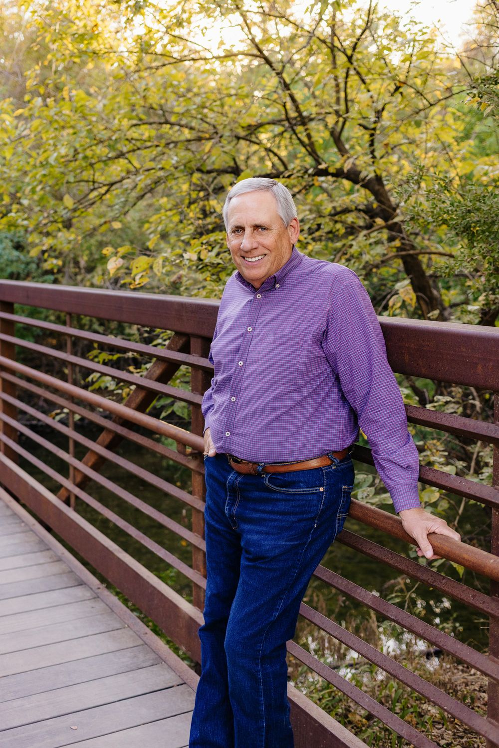 Someone in a purple checkered shirt and jeans stands casually on a wooden bridge with autumn foliage in the background.