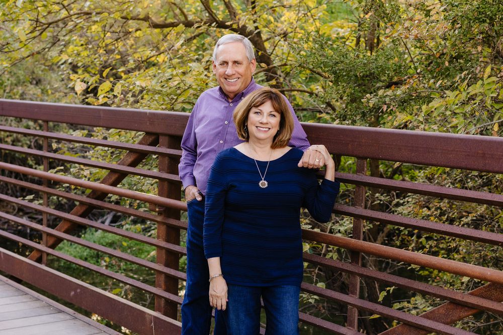 A couple stands together on a wooden footbridge during autumn with yellow foliage in the background.